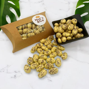 Amandes Stracciatella posées sur une table, accompagnées d'une boîte NutsandCo avec une étiquette, contenant des amandes Stracciatella. À droite, un petit bol noir rempli d’amandes, avec des plantes en arrière-plan.