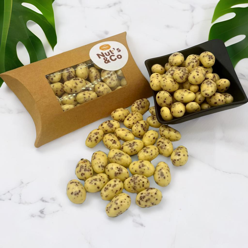 Amandes Stracciatella posées sur une table, accompagnées d'une boîte NutsandCo avec une étiquette, contenant des amandes Stracciatella. À droite, un petit bol noir rempli d’amandes, avec des plantes en arrière-plan.