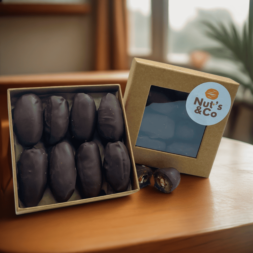 Boîte de Medjoul d'Afrique du Sud au chocolat et amande grillée avec une étiquette NutsAndCo, contenant des Medjoul au chocolat et amande grillée, avec une datte étalée par terre.