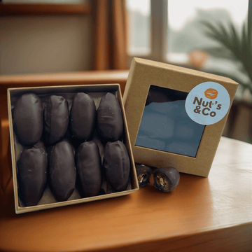 Boîte de Medjoul d'Afrique du Sud au chocolat et amande grillée avec une étiquette NutsAndCo, contenant des Medjoul au chocolat et amande grillée, avec une datte étalée par terre.