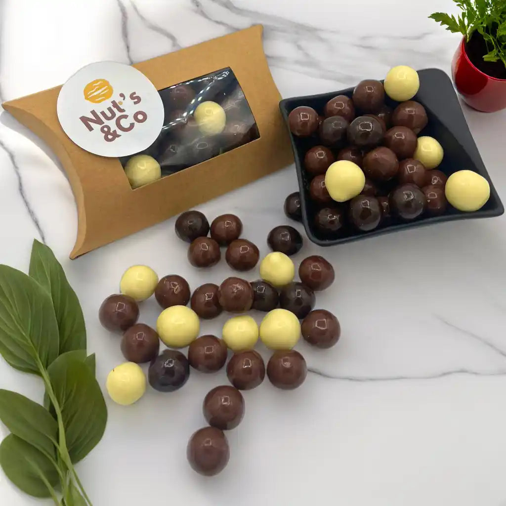 Mix noisette chocolat posé sur une table, accompagné d'une boîte NutsAndCo avec une étiquette, contenant un assortiment de noisettes et de chocolat. À droite, un petit bol noir rempli du mix, avec des plantes en arrière-plan.