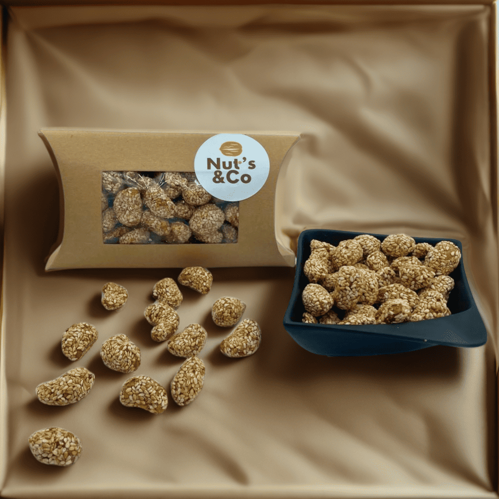 Bol de noix de cajou caramélisées au sésame à côté d’un paquet avec une étiquette NutsAndCo contenant des noix de cajou caramélisées au sésame, avec des noix de cajou étalées par terre.