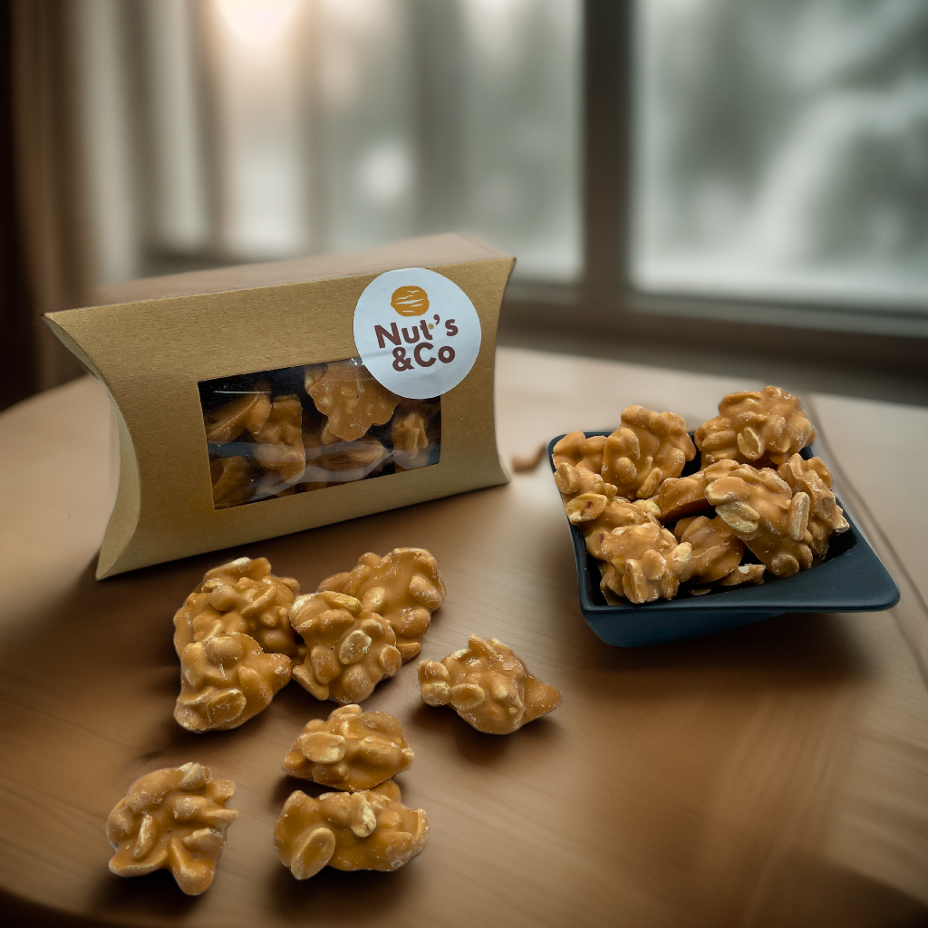 Bol de Rocher Cacahuète Caramel Salé à côté d’une boîte avec une étiquette NutsAndCo contenant des rochers cacahuète caramel salé, avec quelques rochers étalés par terre.